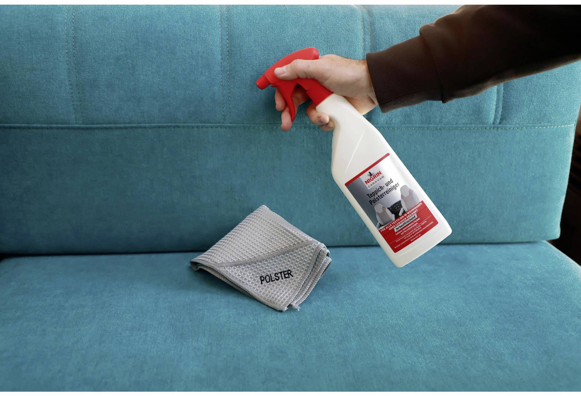 A hand sprays cleaning solution from a bottle onto a turquoise sofa. A cloth with the label 'Upholstery' lies beside it.