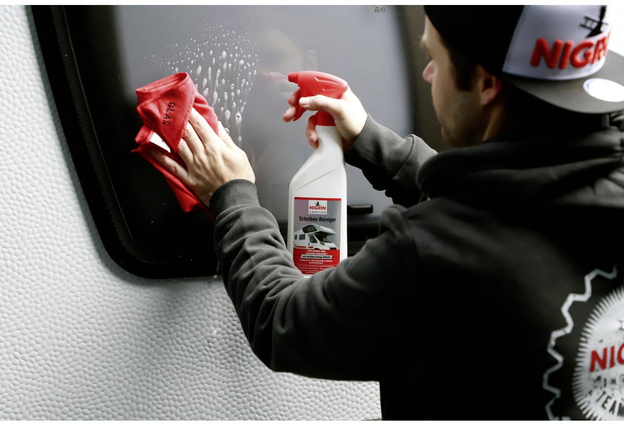 A person is cleaning a window with a spray and a red cloth. The bottle has a label with an image of a car.