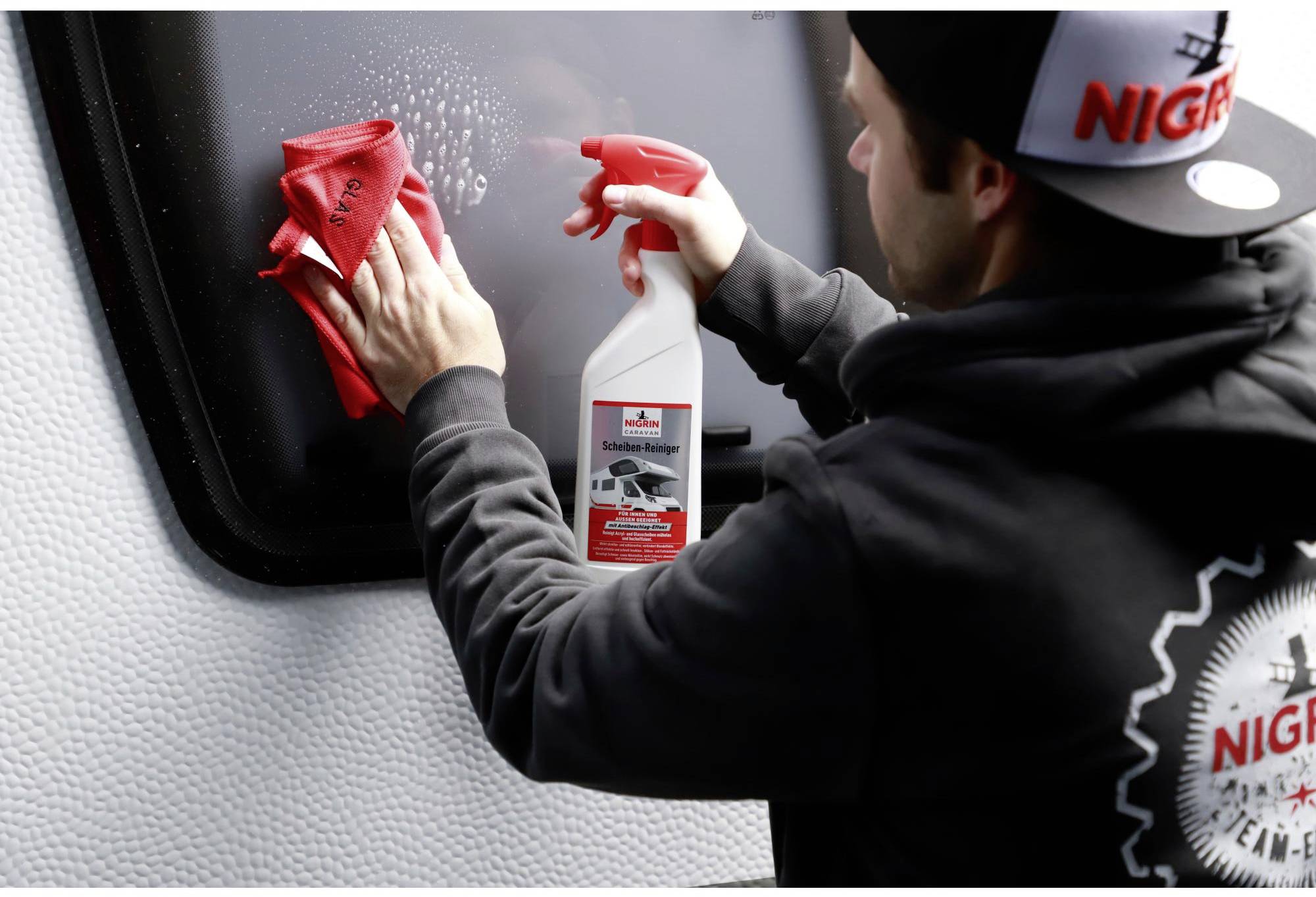 A man is cleaning a dark window with a red cloth and cleaning solution from a spray bottle.