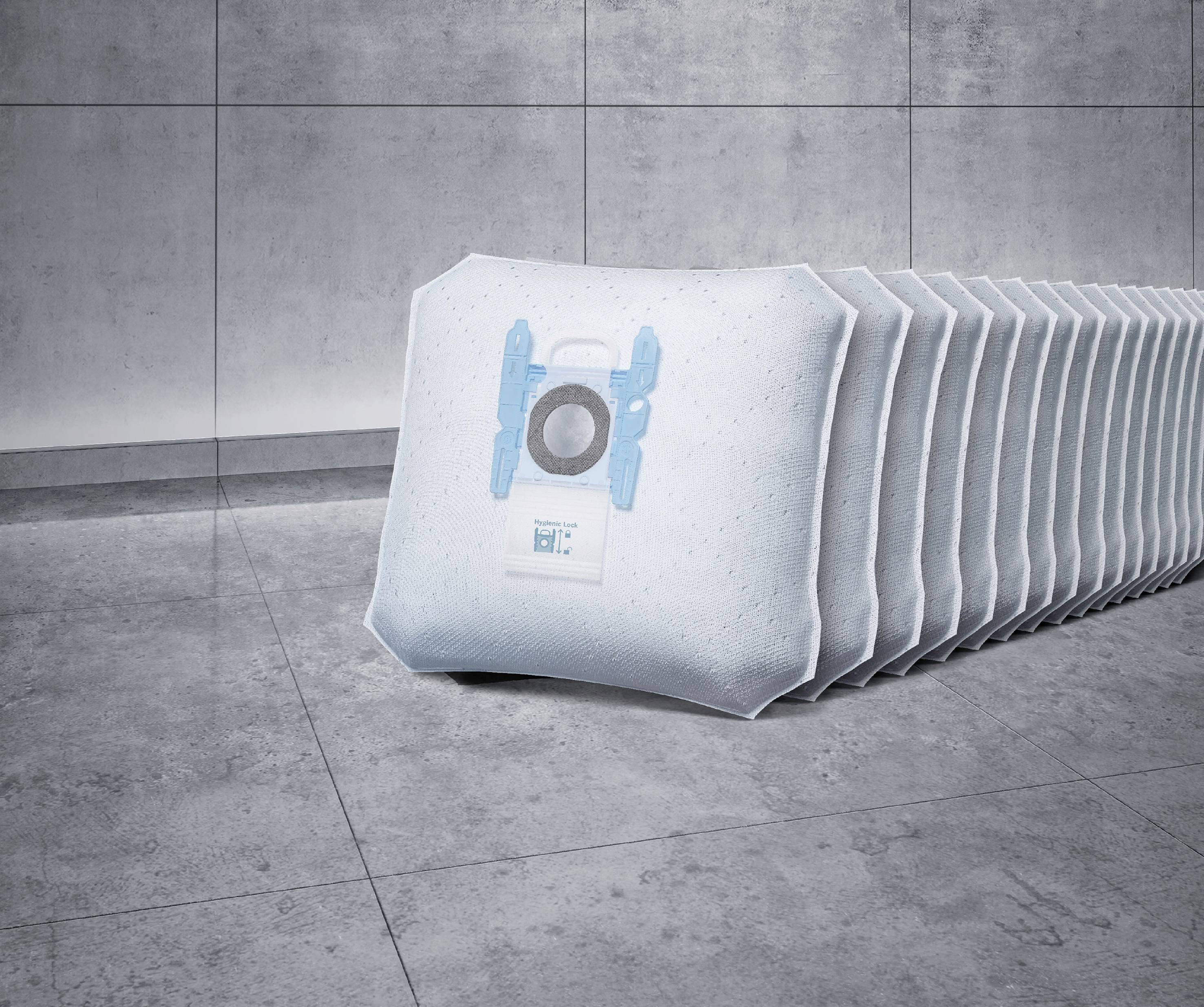 Several white vacuum cleaner bags are lined up in a row on a grey floor. The one at the front shows a round, blue valve.