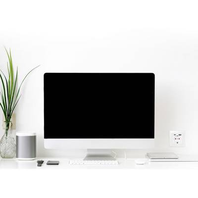 A modern desk with a large computer monitor. Next to it are a plant, a speaker, and a laptop.