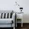 A grey sofa with two patterned cushions sits beside a white table, on which a lamp and a charging smartphone rest.