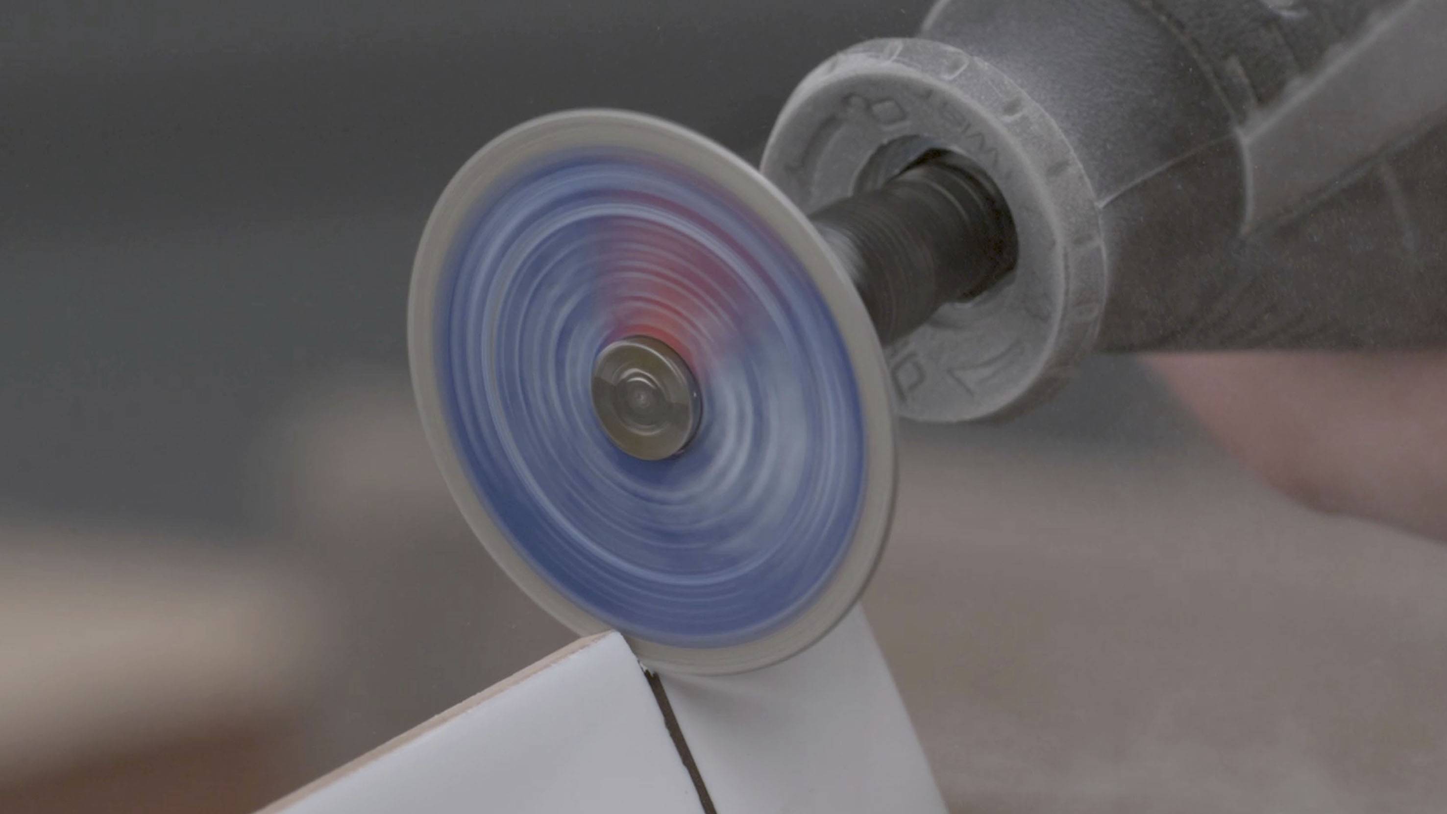 A rotating tool grinds a white surface, while dust billows in the background.