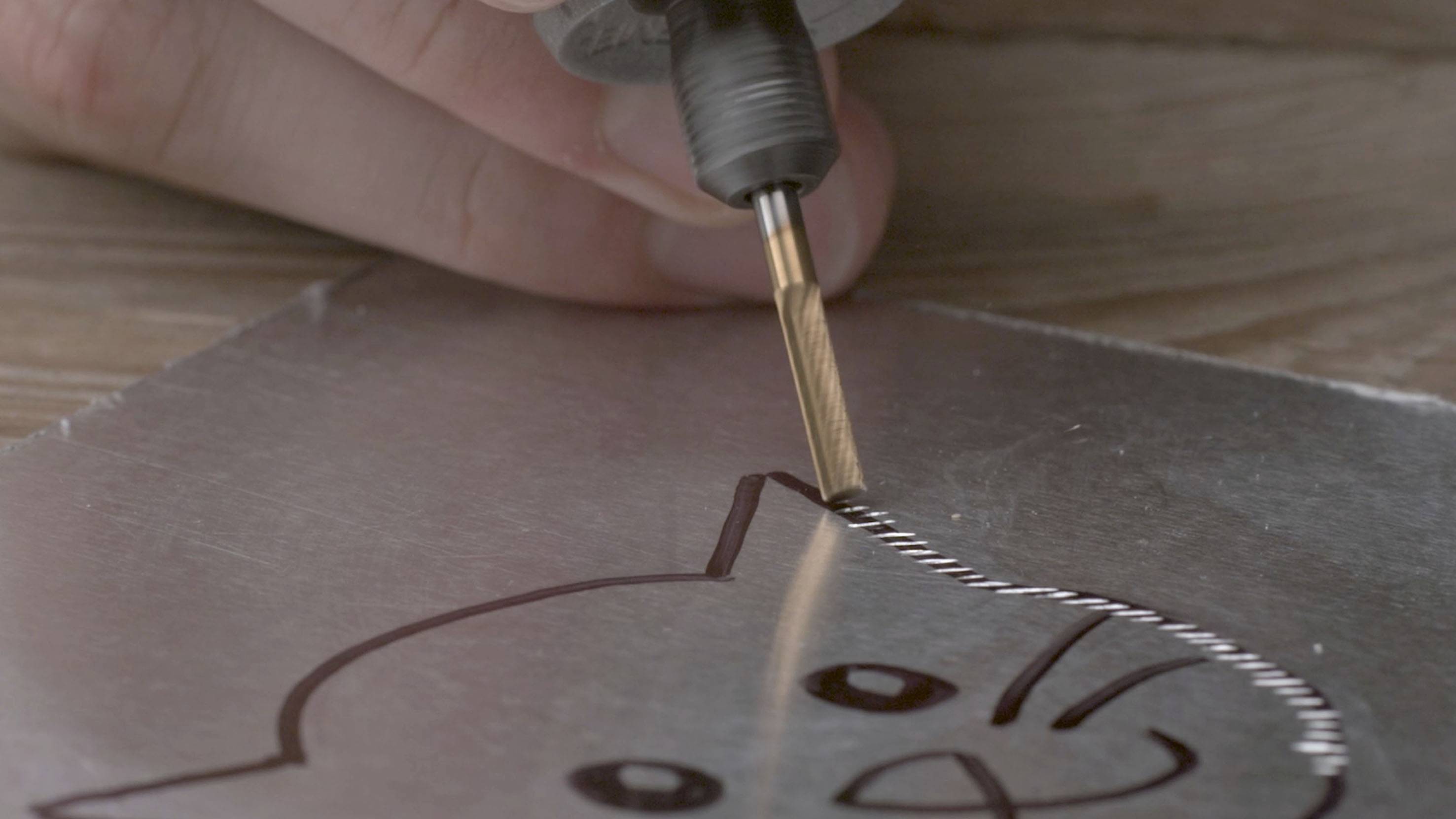 A close-up shows a hand using a metal tool to engrave the outline of a cat onto a metal plate.