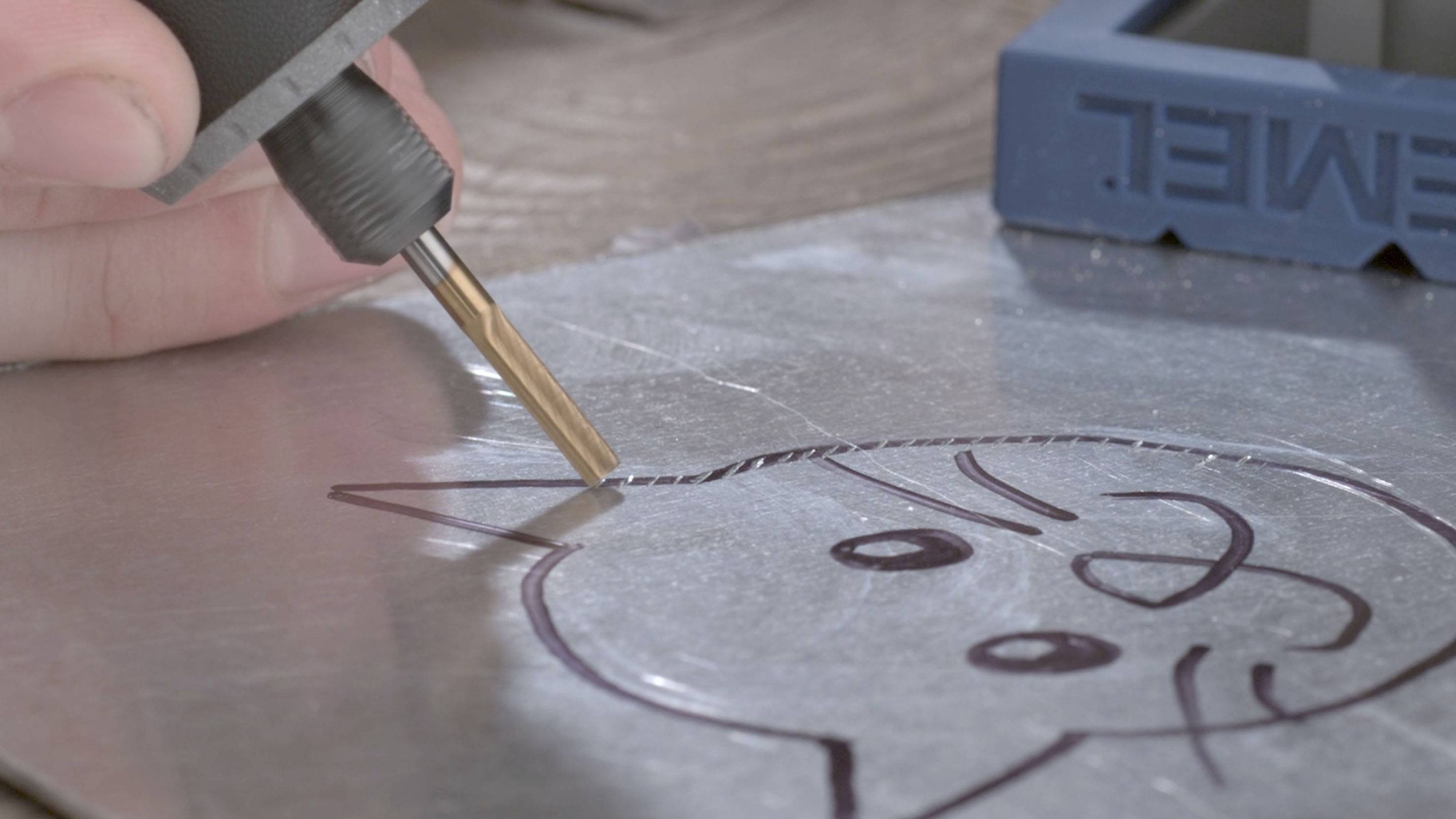A hand holds an engraving tool and works on a metal plate with a drawn cat sketch on it.