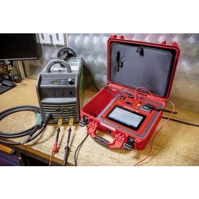 A red electronic testing device connected via cables to another device on a workbench. A metal wall is visible in the background.