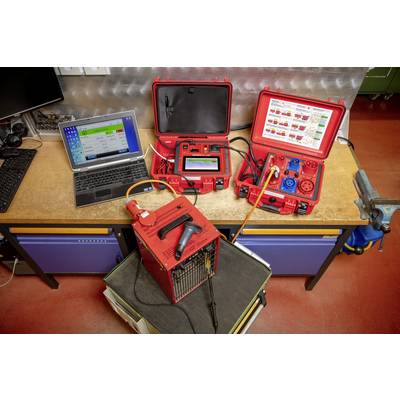 Two red suitcases with technical equipment are standing on a workbench next to a laptop. A device is testing electrical connections.