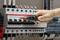 A hand with a voltage tester touches a fuse box to check the voltage. Cables lead into the box.