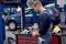 A mechanic in a workshop is examining an electronic device next to a mobile tool trolley, while a car is raised on a lift.