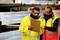 Two men in protective clothing and respirator masks are checking a clipboard in an industrial setting with metal shelving in the background.