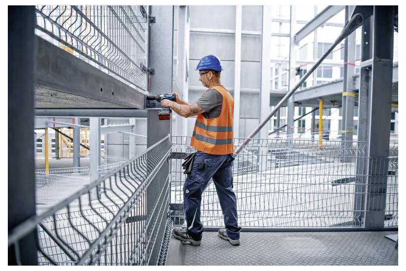 A construction worker wearing a blue helmet and orange vest uses a power tool on a metal frame at a building site.