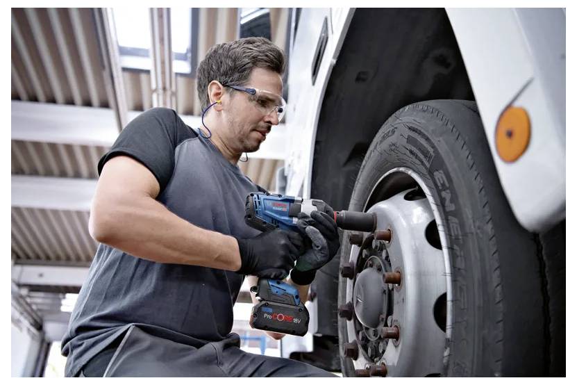 A person wearing safety gear uses a power tool to remove a wheel from a large vehicle in a garage setting.