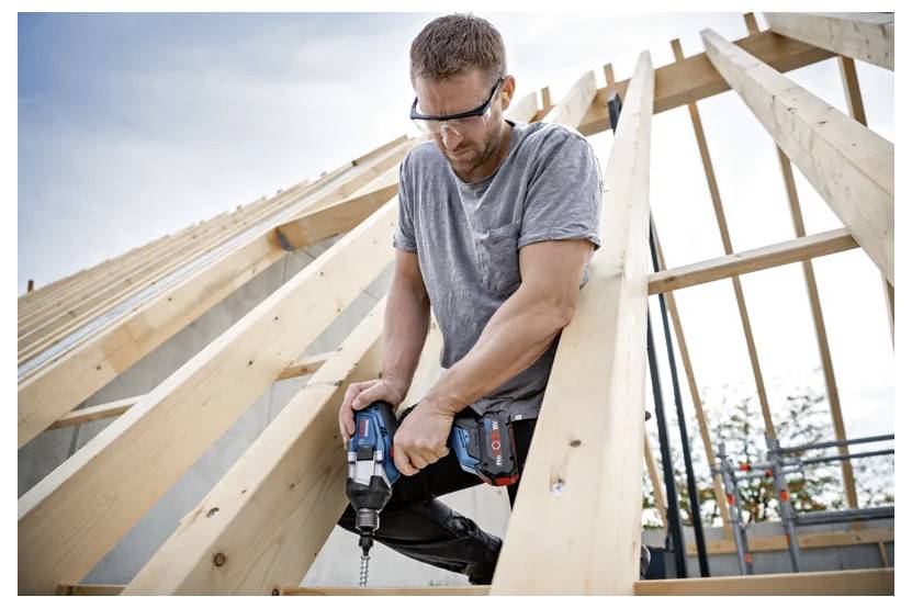 A person wearing safety glasses uses a power drill on wooden beams at a construction site, highlighting carpentry work in progress outdoors.