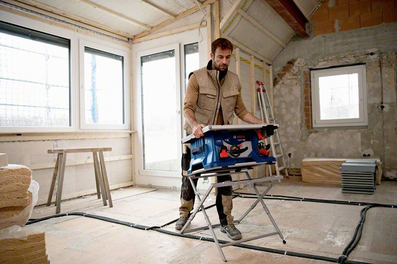 A worker in work attire is using tools on a construction site. Unfinished walls are in the background, and the site appears active and bustling.