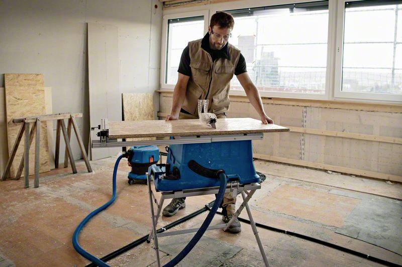 A person is working on a wooden board using a table saw in an unfinished workshop with large windows.