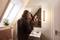 A person is smoothing her hair in the bathroom in front of a mirror. A light is on the right, and a plant is standing on the windowsill.