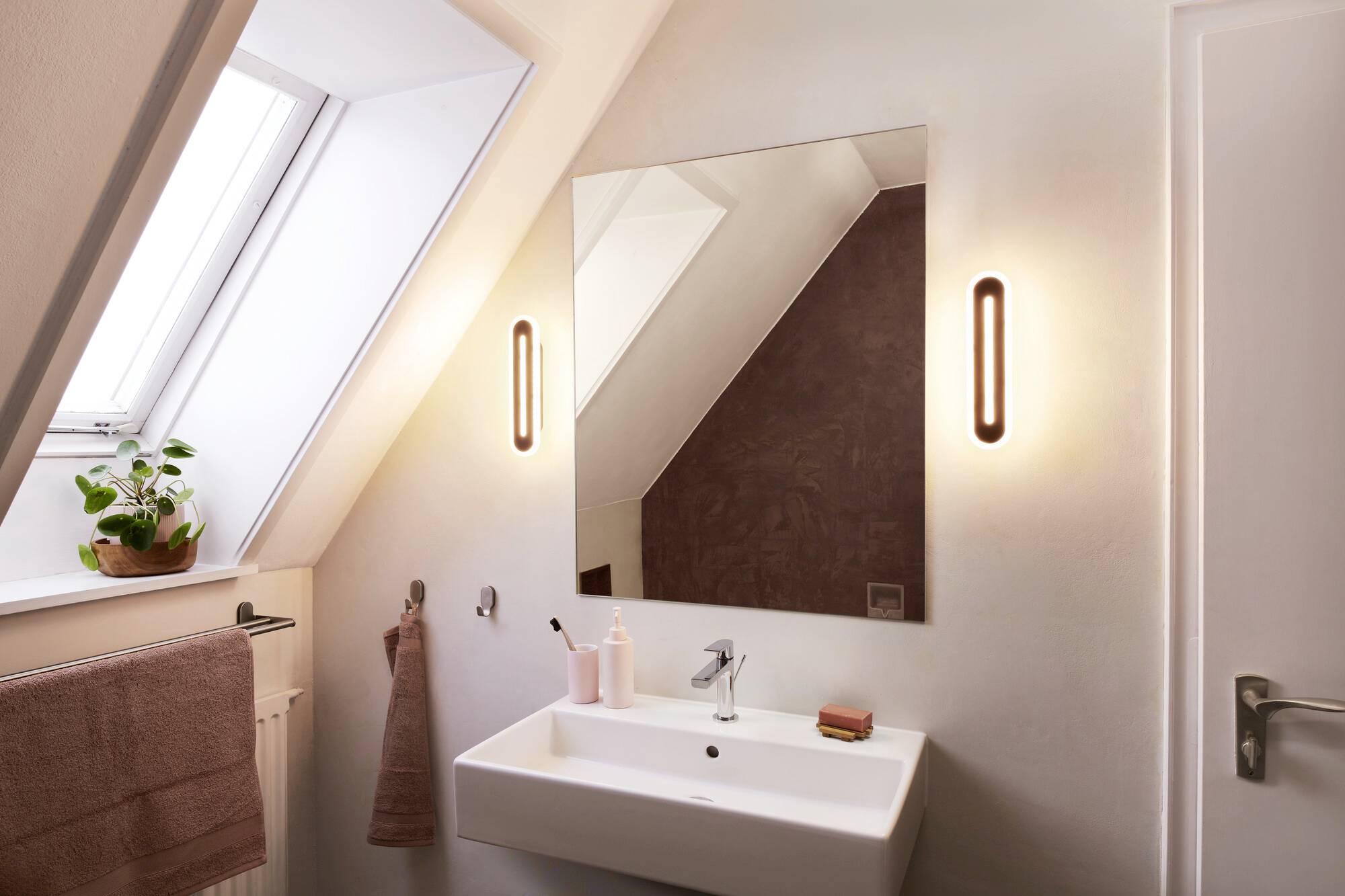 Small, modern bathroom with a roof window. A rectangular mirror hangs above a white washbasin. Lighting on the sides.