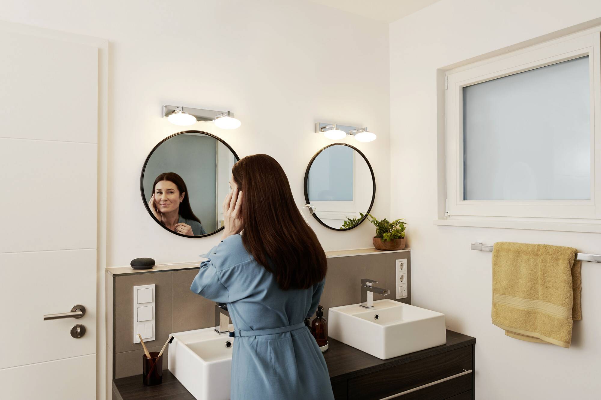 A woman stands in a bathroom in front of a double washbasin, looking into the mirror. She is wearing a blue dressing gown.