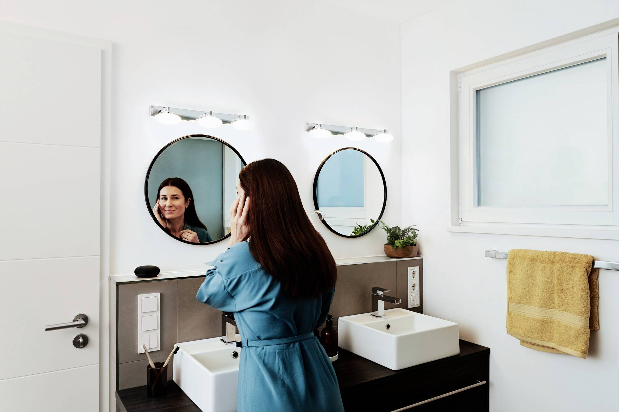 Woman in a blue dress looks in the mirror in the bathroom. Two round mirrors, two washbasins, a plant and a towel in the background.