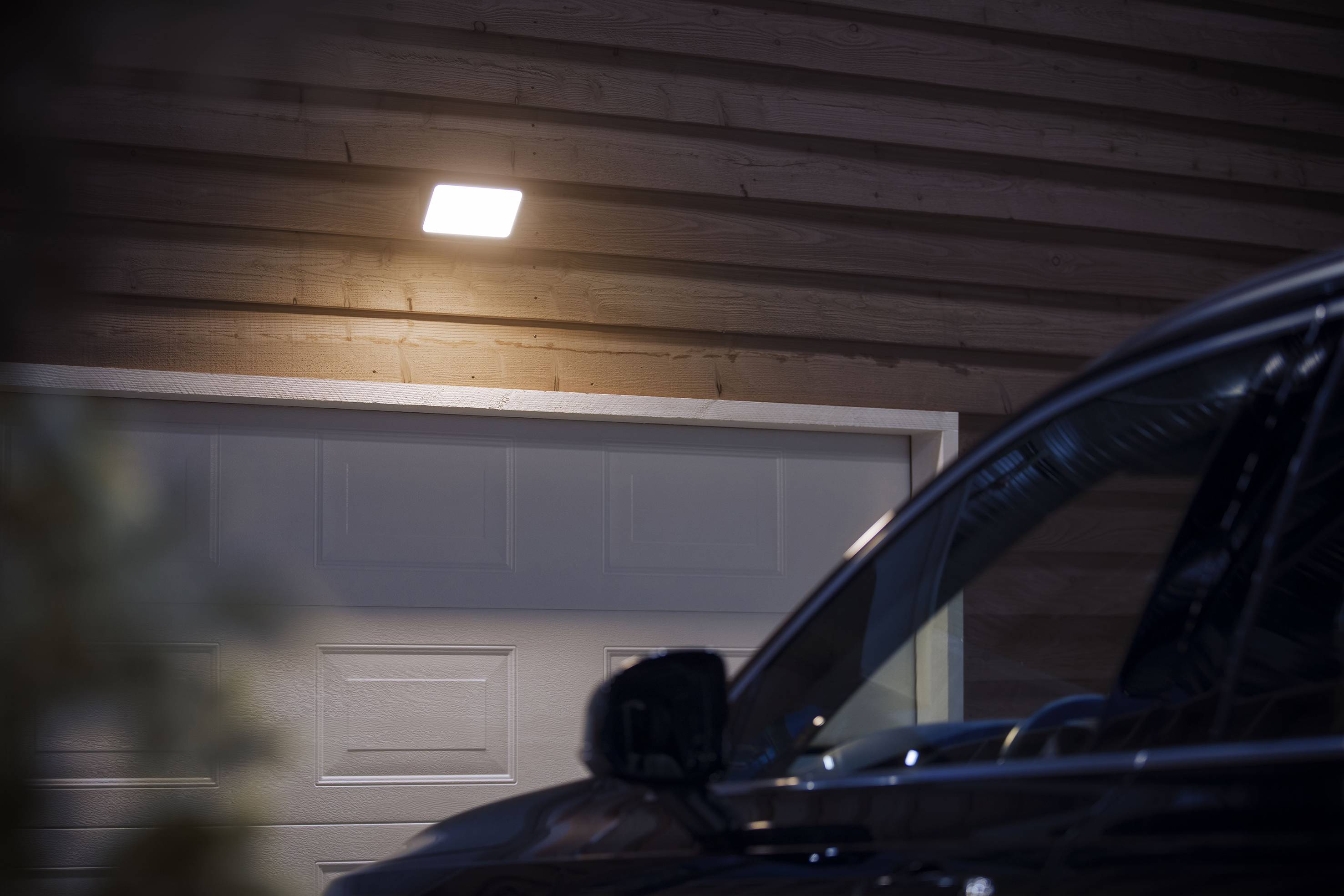 A car is parked in front of a closed garage door, illuminated by an exterior light.