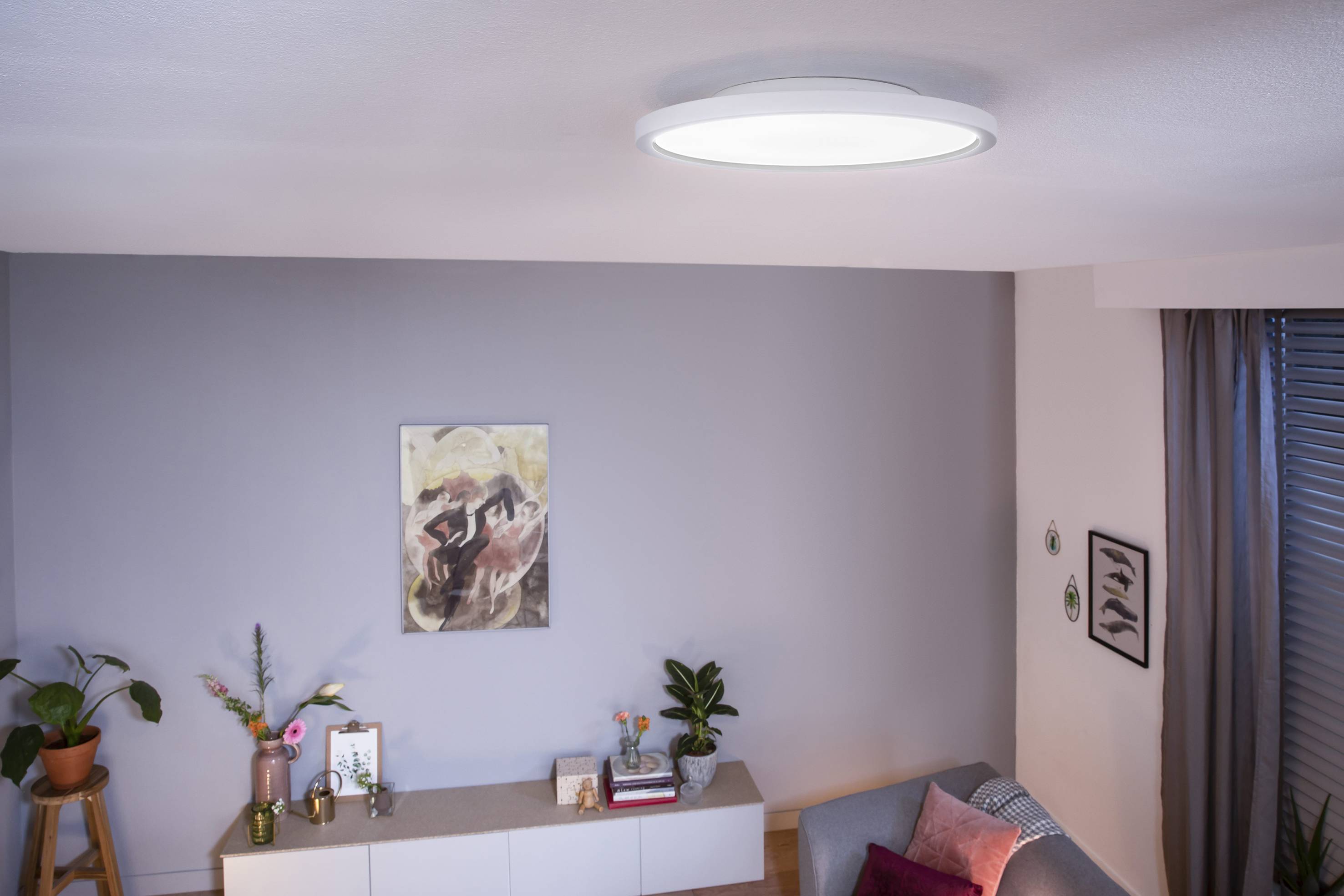Living room with a modern ceiling light, plain white wall, painting, plants, sideboard and grey sofa. Cosy atmosphere.