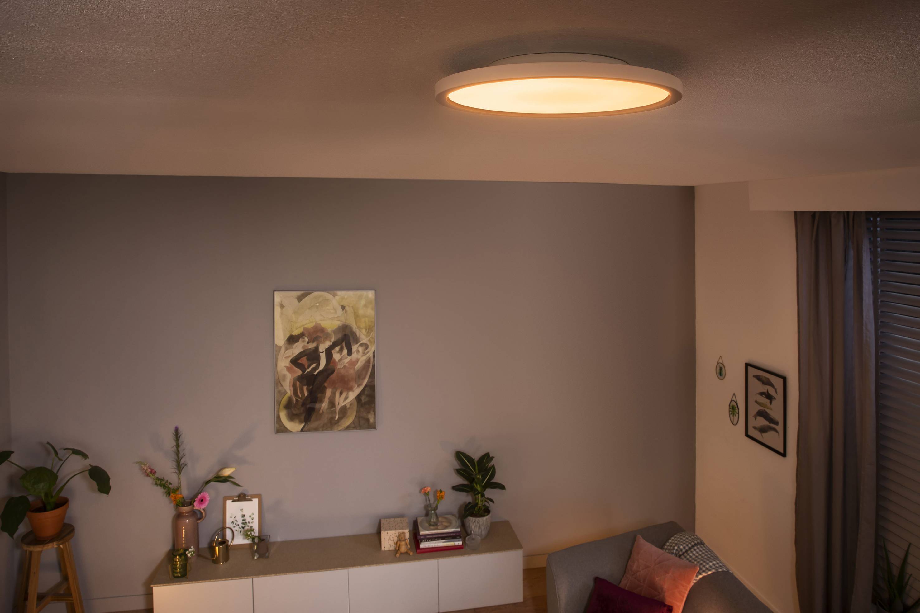 A living room with a round, illuminated ceiling light. Beneath it, a sideboard with plants and pictures, a sofa with cushions.