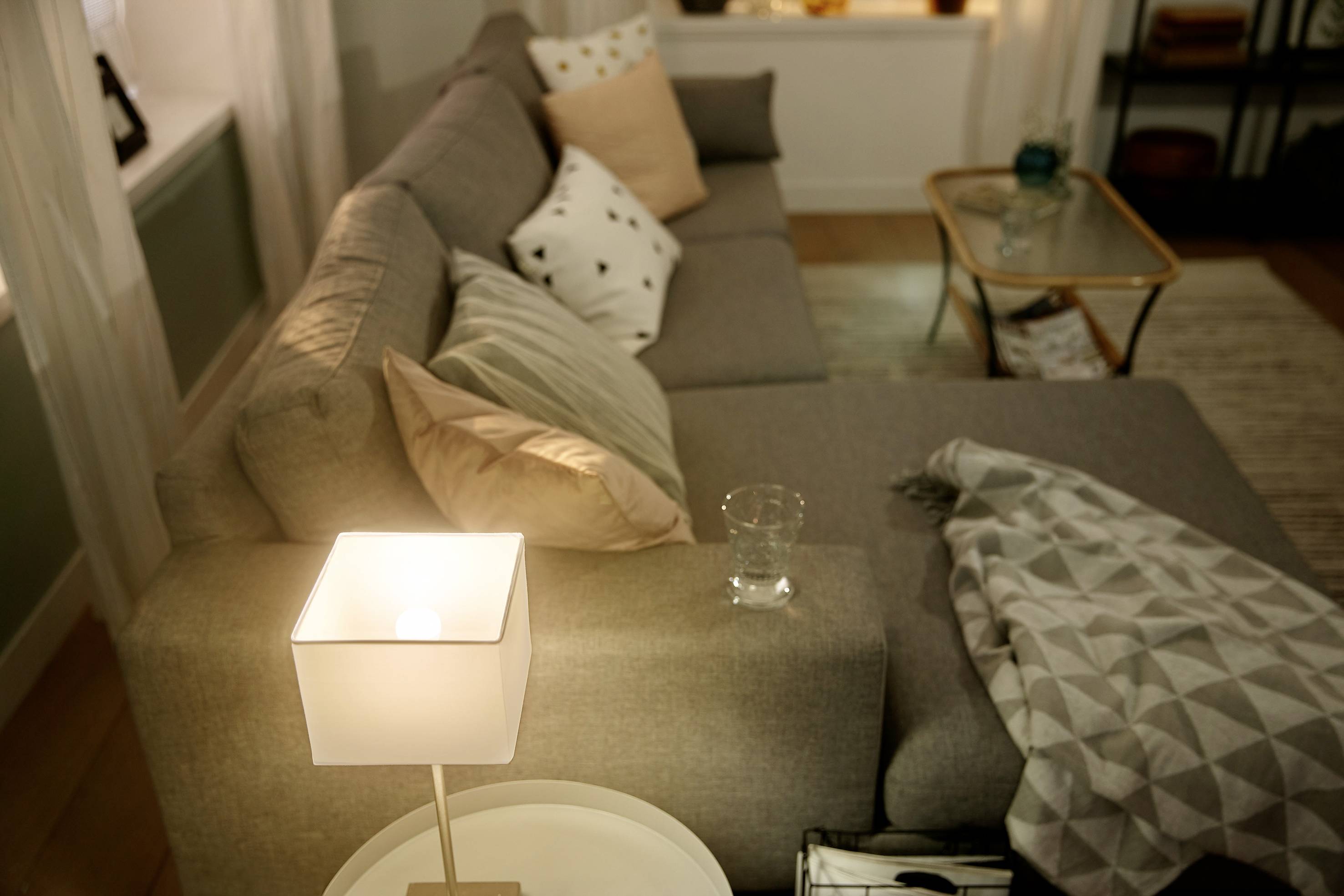 Cosy living room with a grey sofa, decorated with cushions and a throw. A glowing table lamp sits on a small side table.