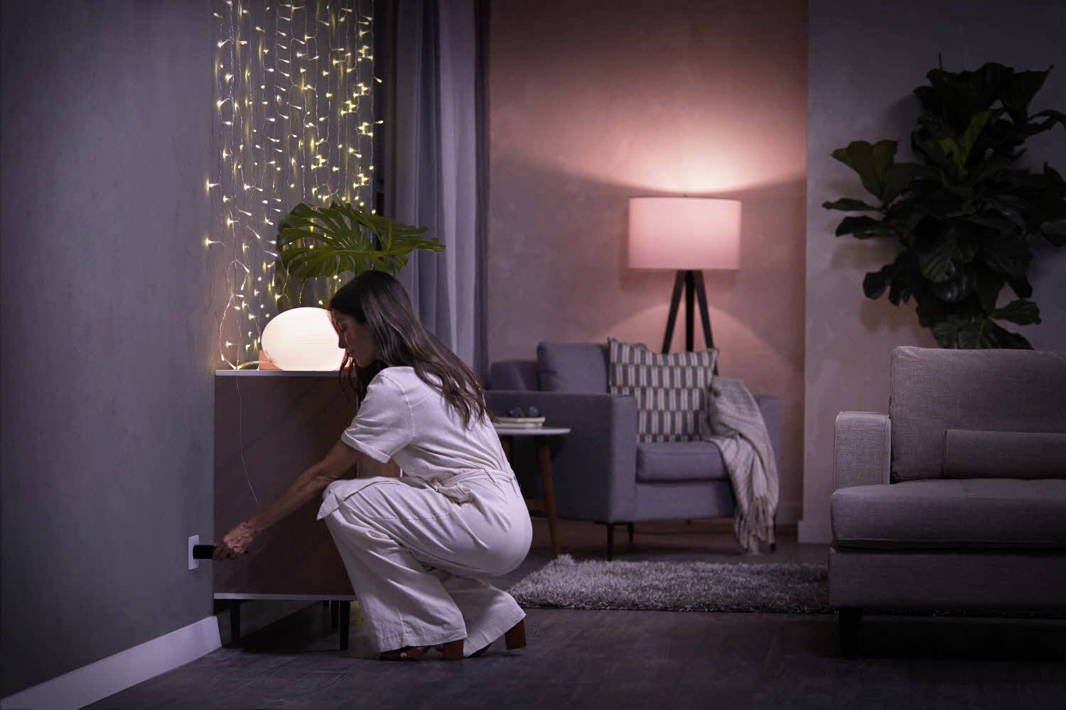 A woman in light-coloured clothing kneels down and plugs something into a socket. The room is cosily lit with plants and furniture.