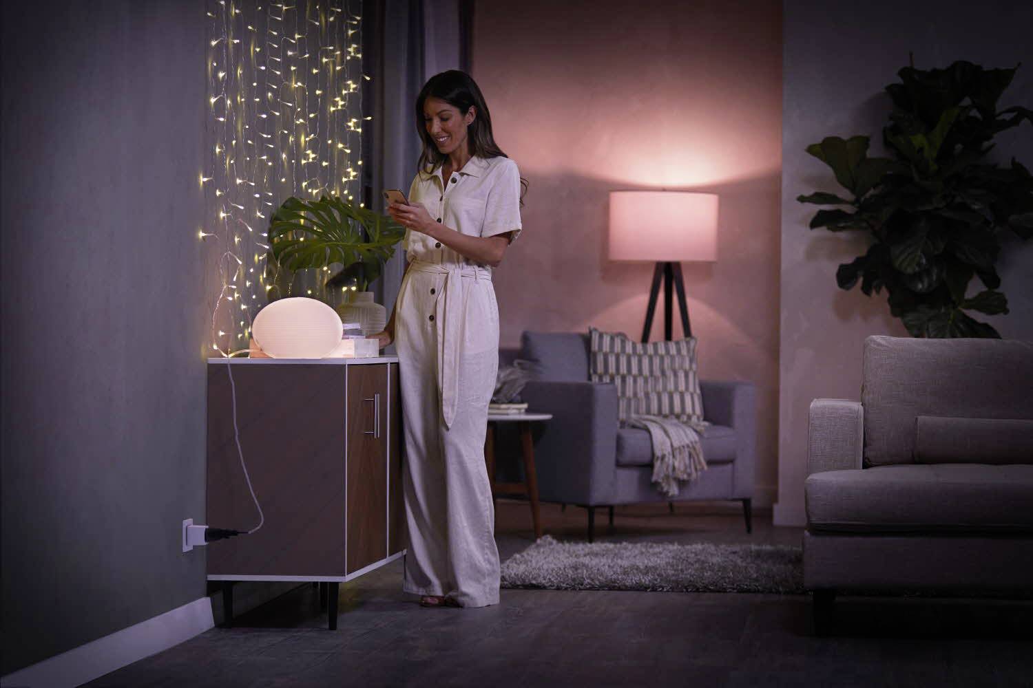 A woman stands beside a table with a decorative lamp and fairy lights in a cosily lit living room, smiling.