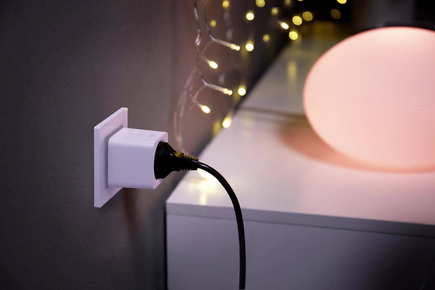 A plug socket with a black cable plugged in. Next to it, a table with a glowing lamp and fairy lights in the background.