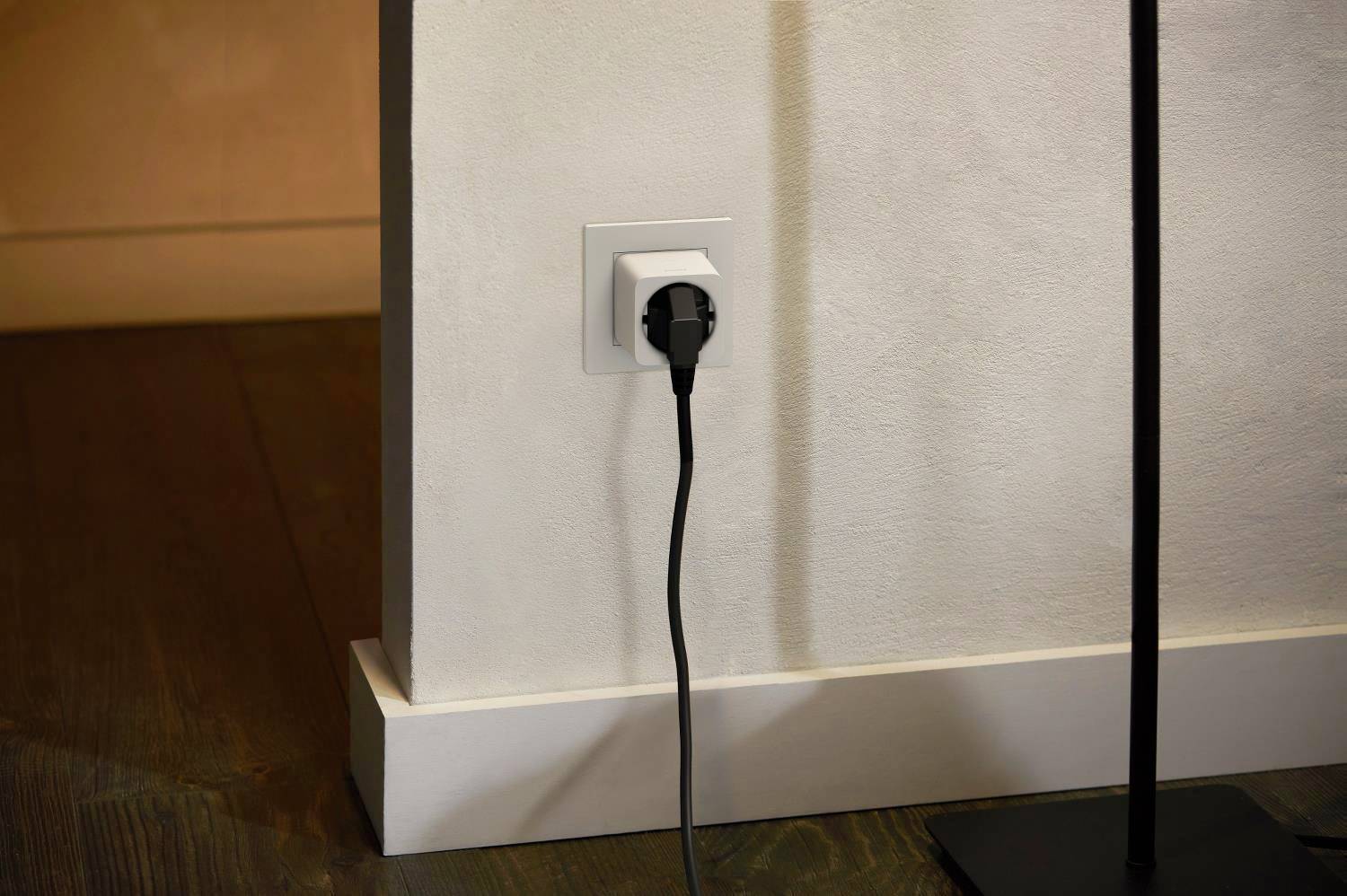 A black socket is mounted on a white wall, with a cable plugged in and leading to the floor. To the left, a wooden floor is visible.