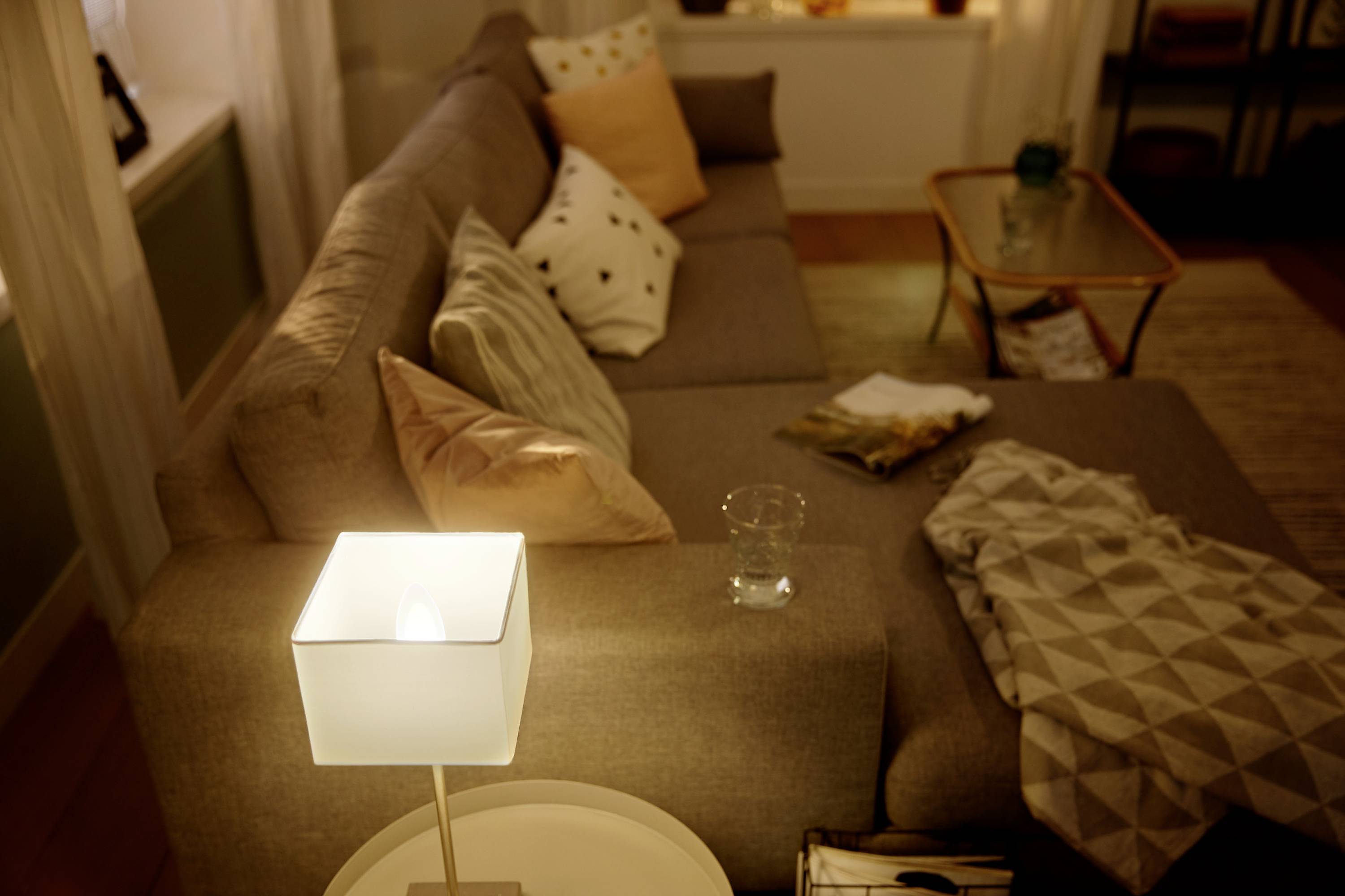 A cosy living room with a large grey sofa, adorned with various cushions and throws, alongside a glowing table lamp.