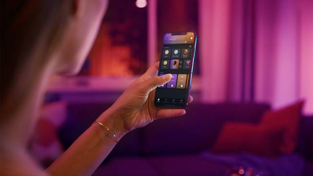 A person is controlling the smart home lighting with a smartphone in a cosy living room with purple illumination.
