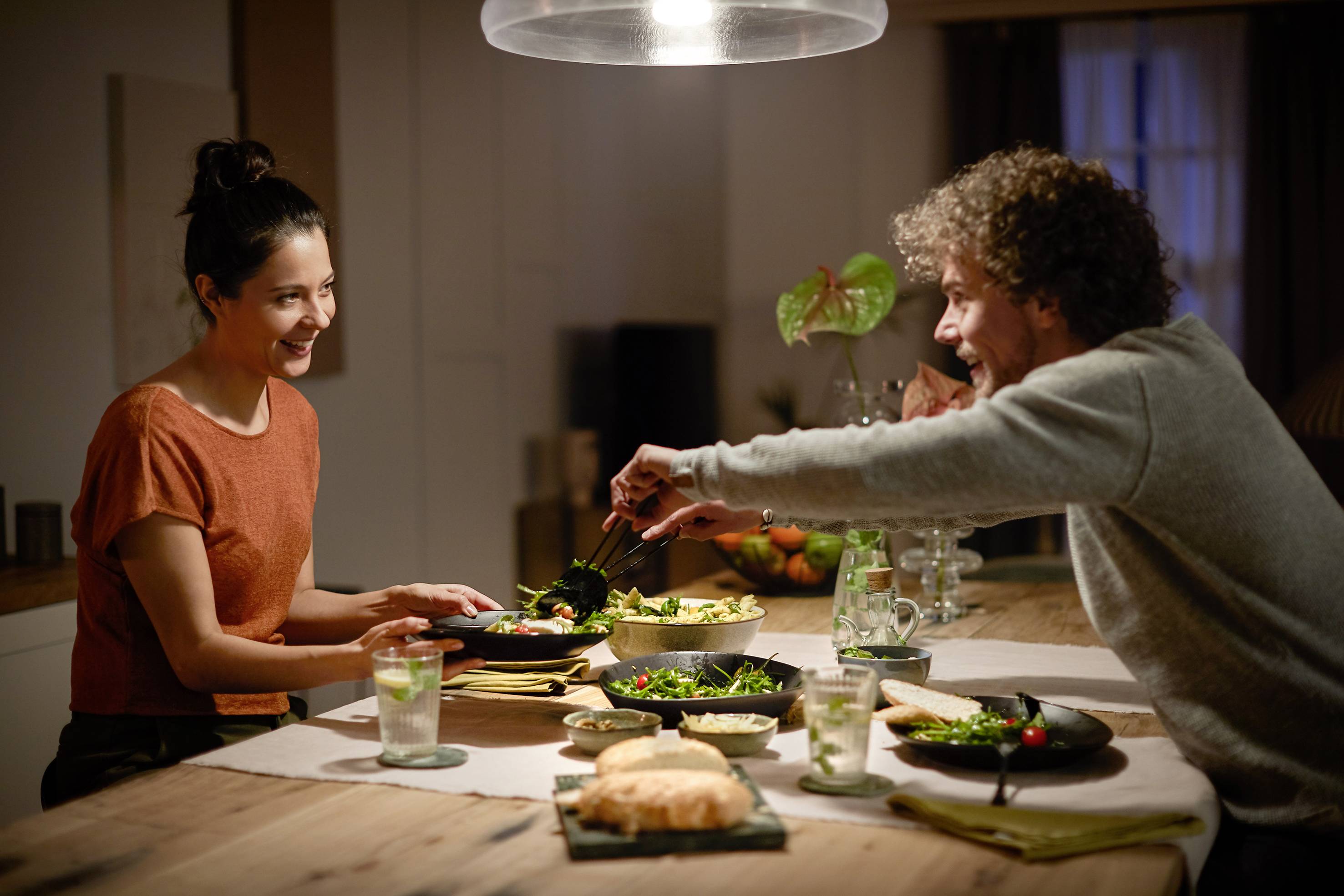 A man and a woman are sitting at a table, laughing and eating together. The table is laid with various dishes.