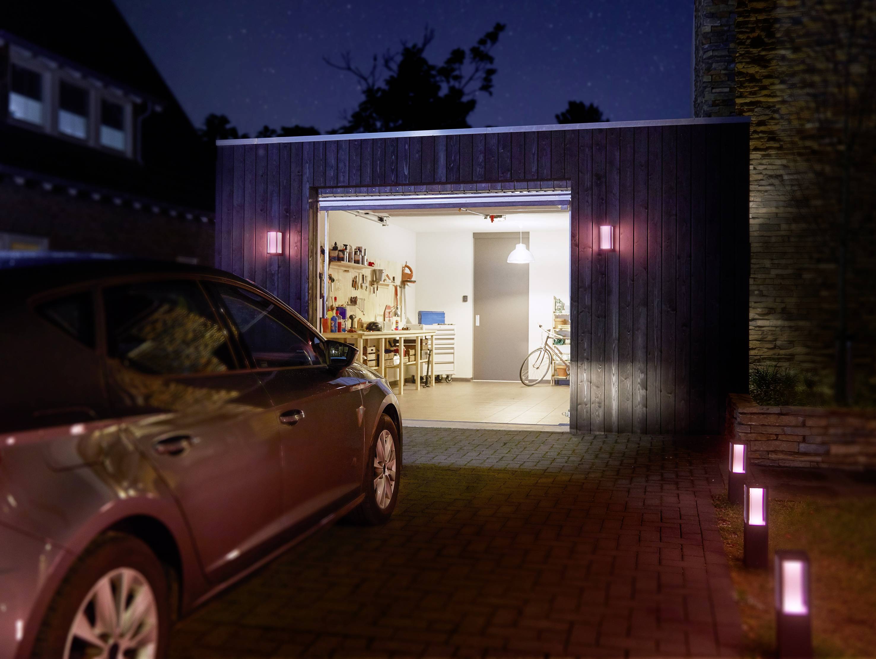 A car is parked in a driveway in front of an open garage. The garage is illuminated and contains tools and a bicycle.