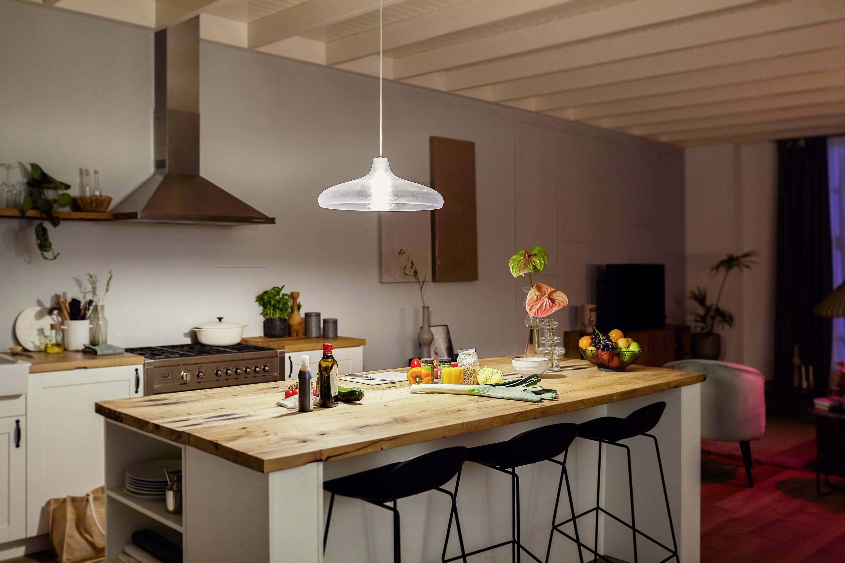 Modern kitchen with a wooden island and three bar stools, with a hanging light above. Fruit, vegetables, and crockery are placed on the island.