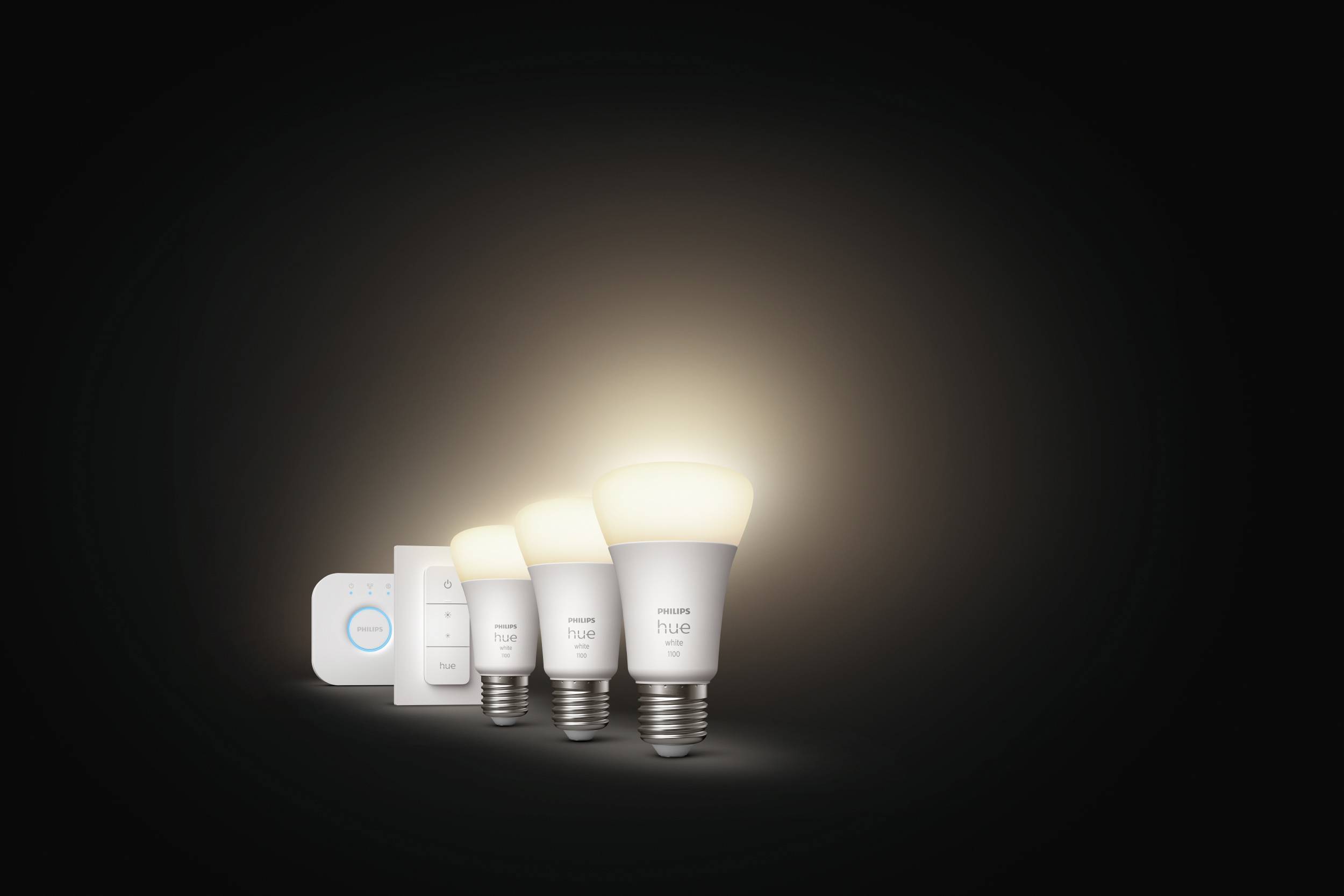 'Three Philips Hue light bulbs and a control unit against a dark background. The light bulbs are glowing softly and displaying their colour variety.'