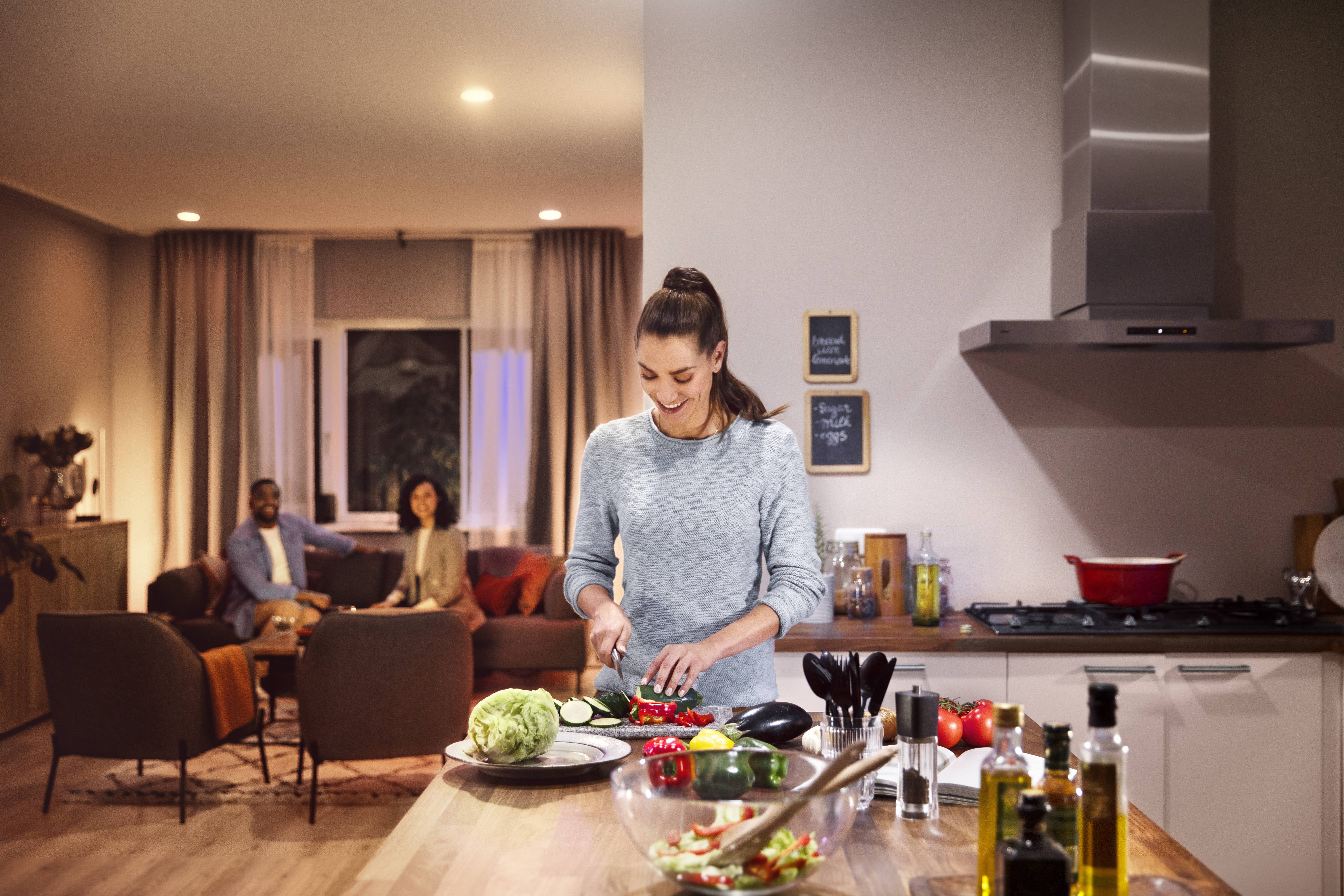 A woman is chopping vegetables in a modern kitchen. In the background, two people are sitting in the living room and chatting.