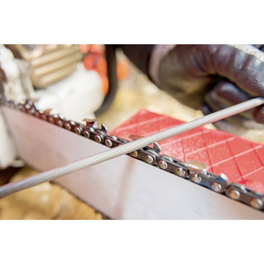 Close-up of a person sharpening a chainsaw with a file, wearing black gloves. Blurred wood in the background.
