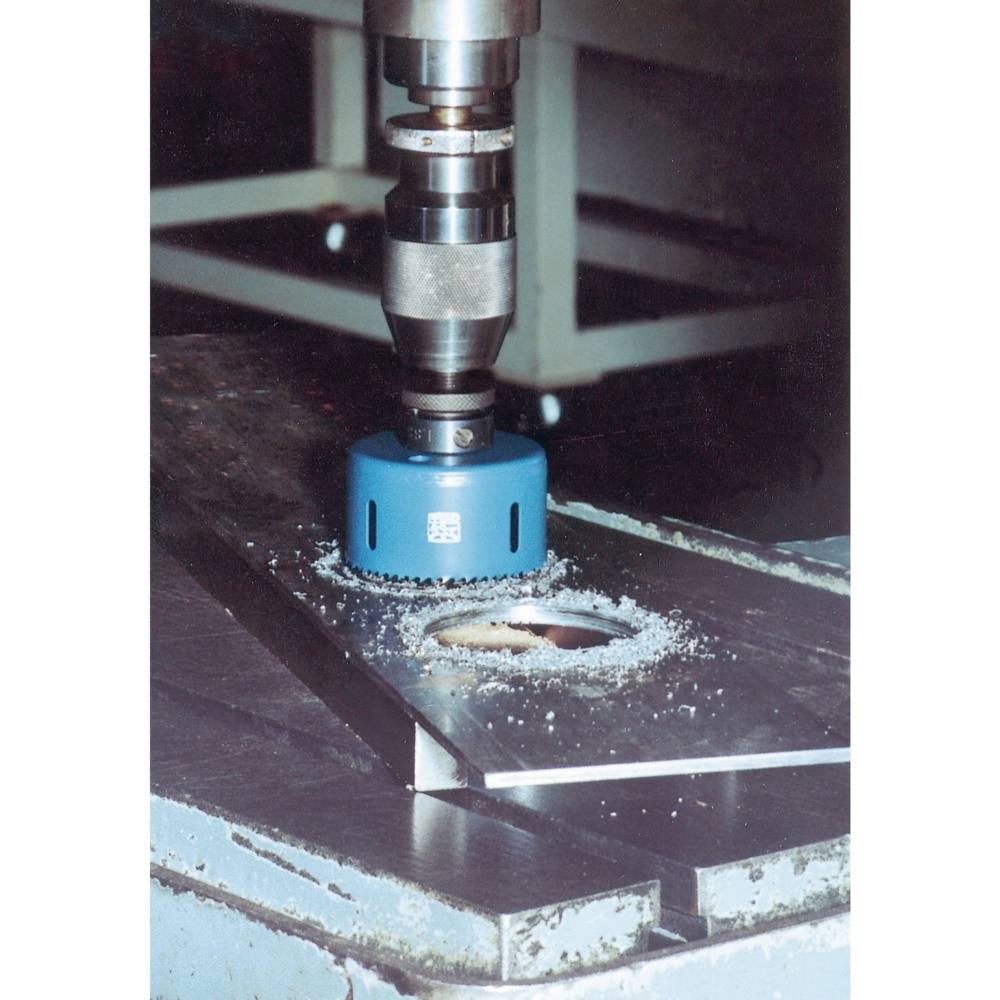 A blue hole saw drills a round hole into a metal plate on a workbench. Metal shavings are visible around the hole.
