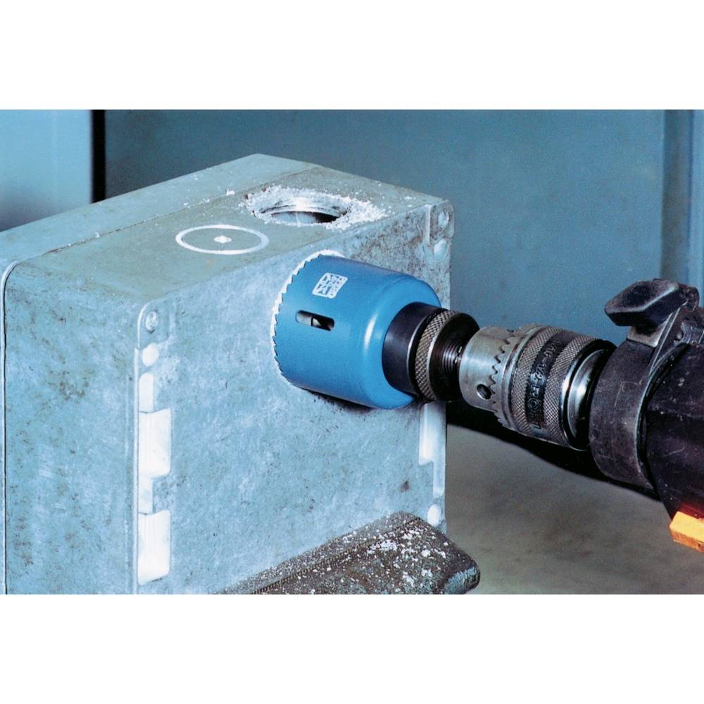 A drill with a hole saw drills a large, round hole in a metal box. Fluff of metal shavings around the hole.