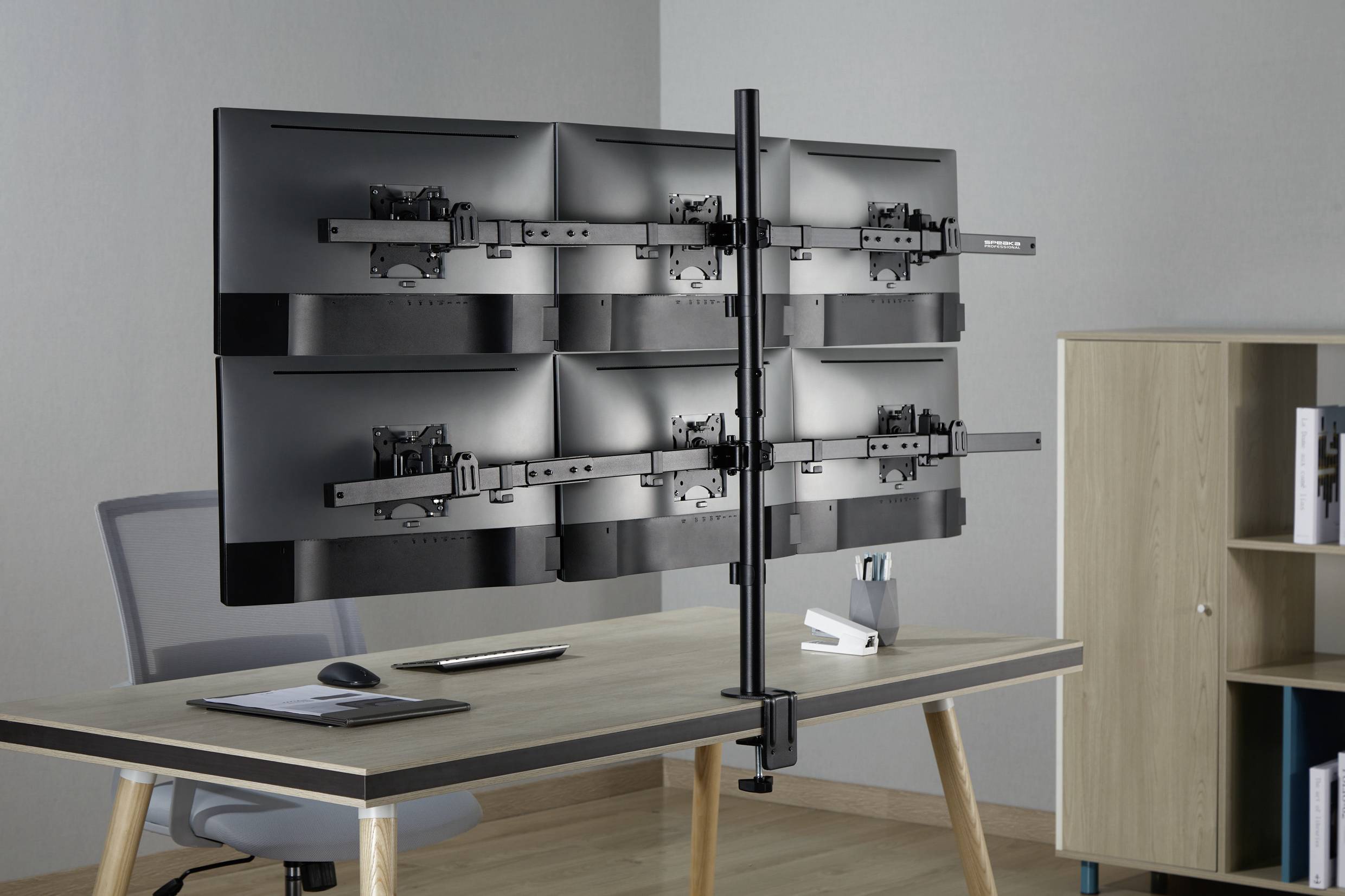 A desk clamp holds six monitor mounts, attached in two stacked rows, on a desk in an office.