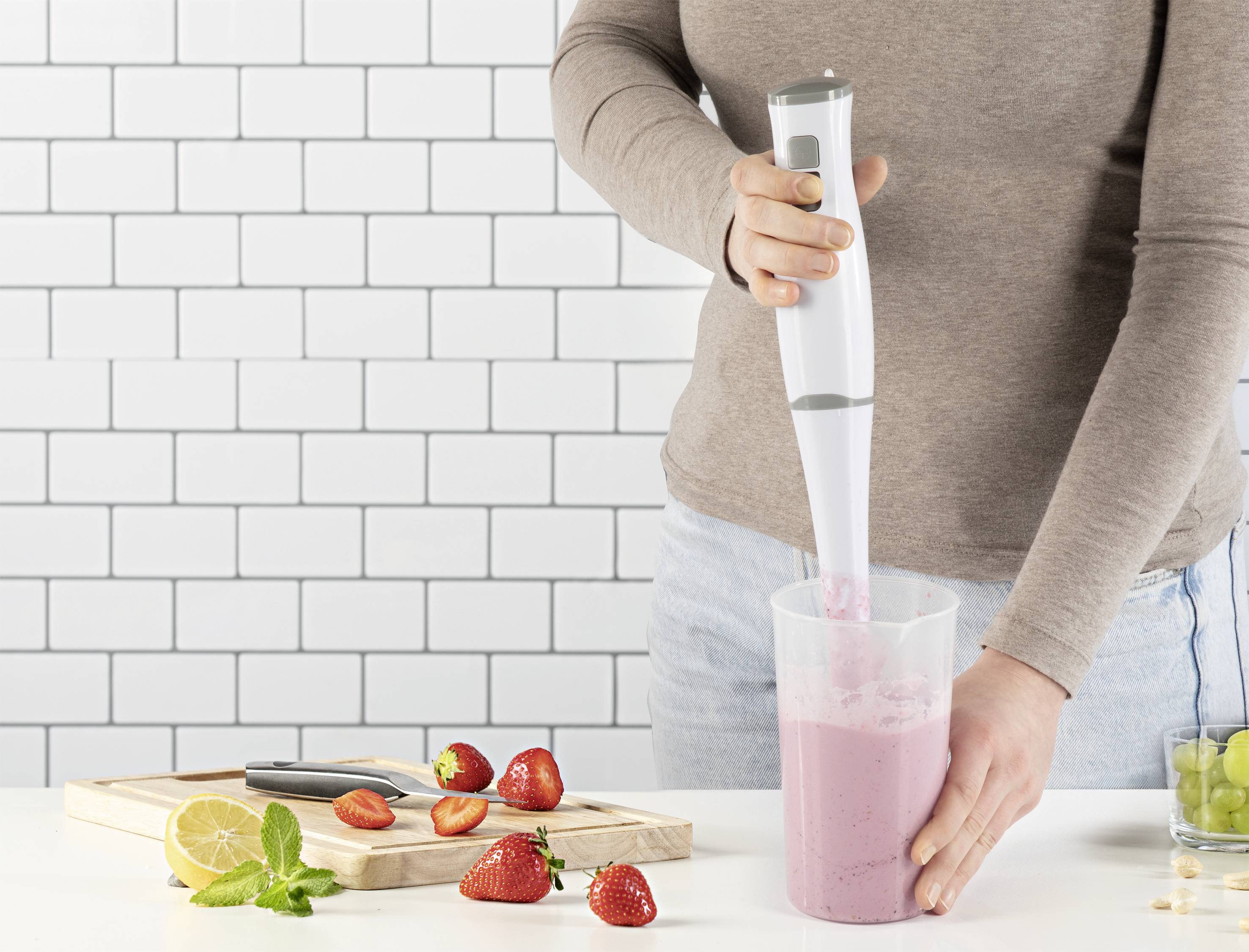 A person is blending a strawberry smoothie with a hand mixer in a measuring jug. Strawberries and lemon slices are lying on the table.