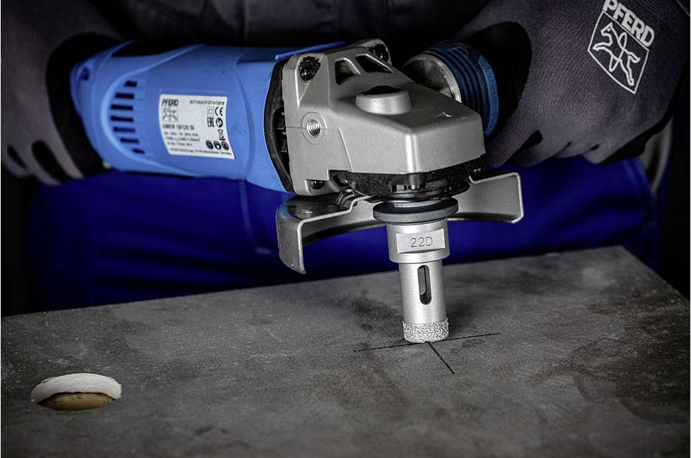 A person is wearing gloves and operating a blue electric drill, creating a hole in a grey surface.