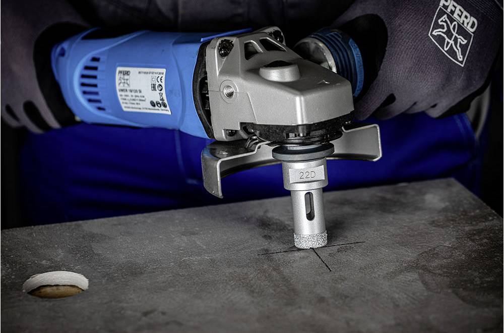 A person is working on a metal plate with an angle grinder in a workshop environment. The plate has a drilled hole.