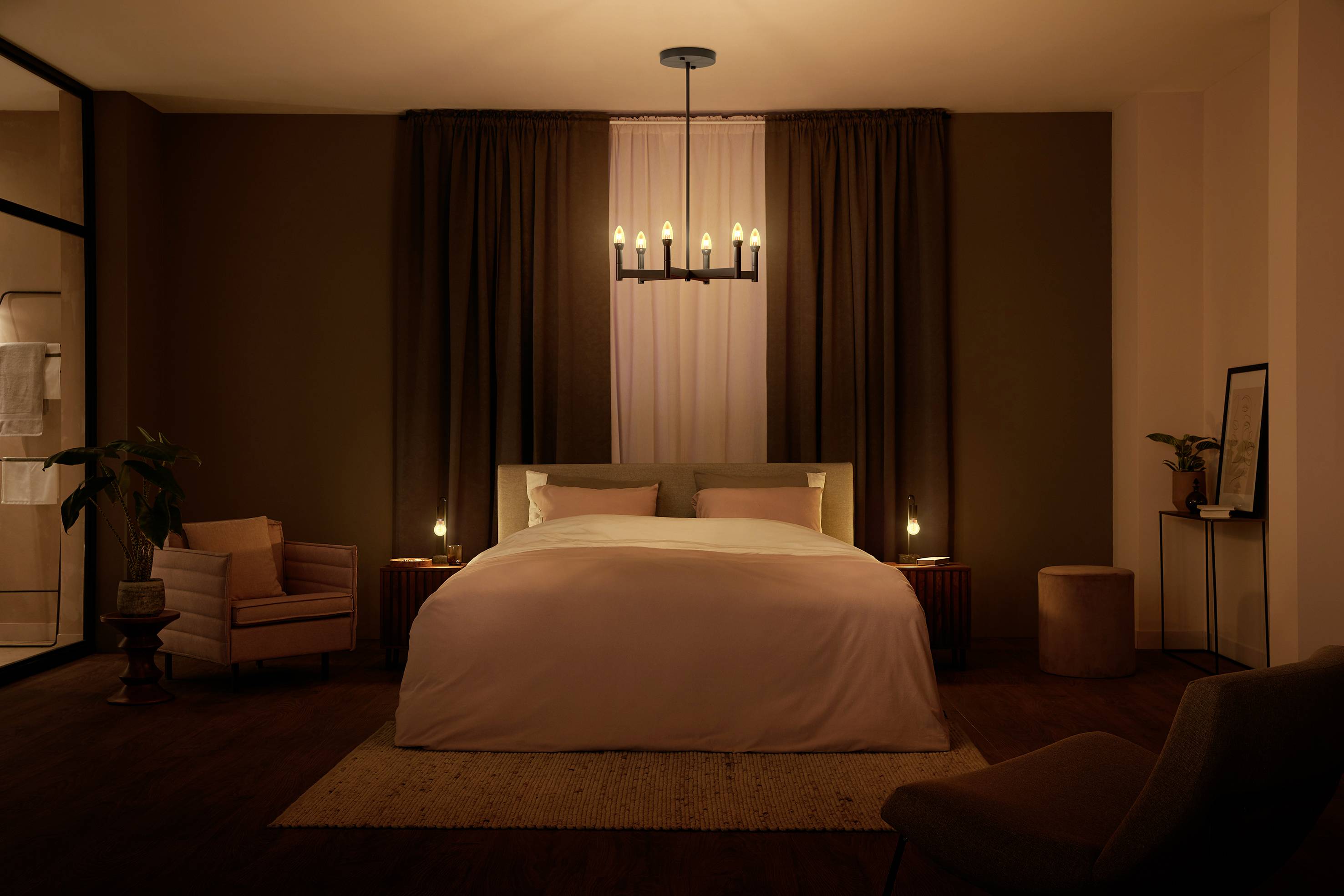 Cosy bedroom with a large bed, warm lighting chandelier, two bedside lamps, an armchair and plants.