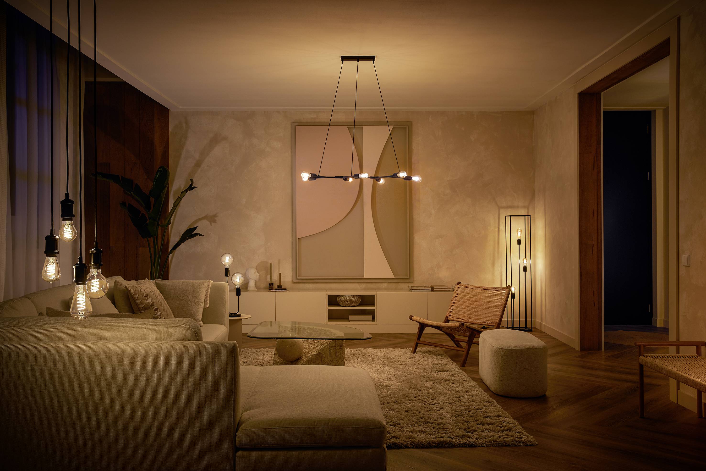 Modern living room with subtle lighting, beige sofa and chair, wooden flooring, rug and large plants. Understated wall art.