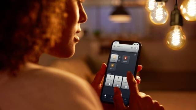 A person is using a smartphone to control smart lighting in a room. Hanging light bulbs can be seen in the background.