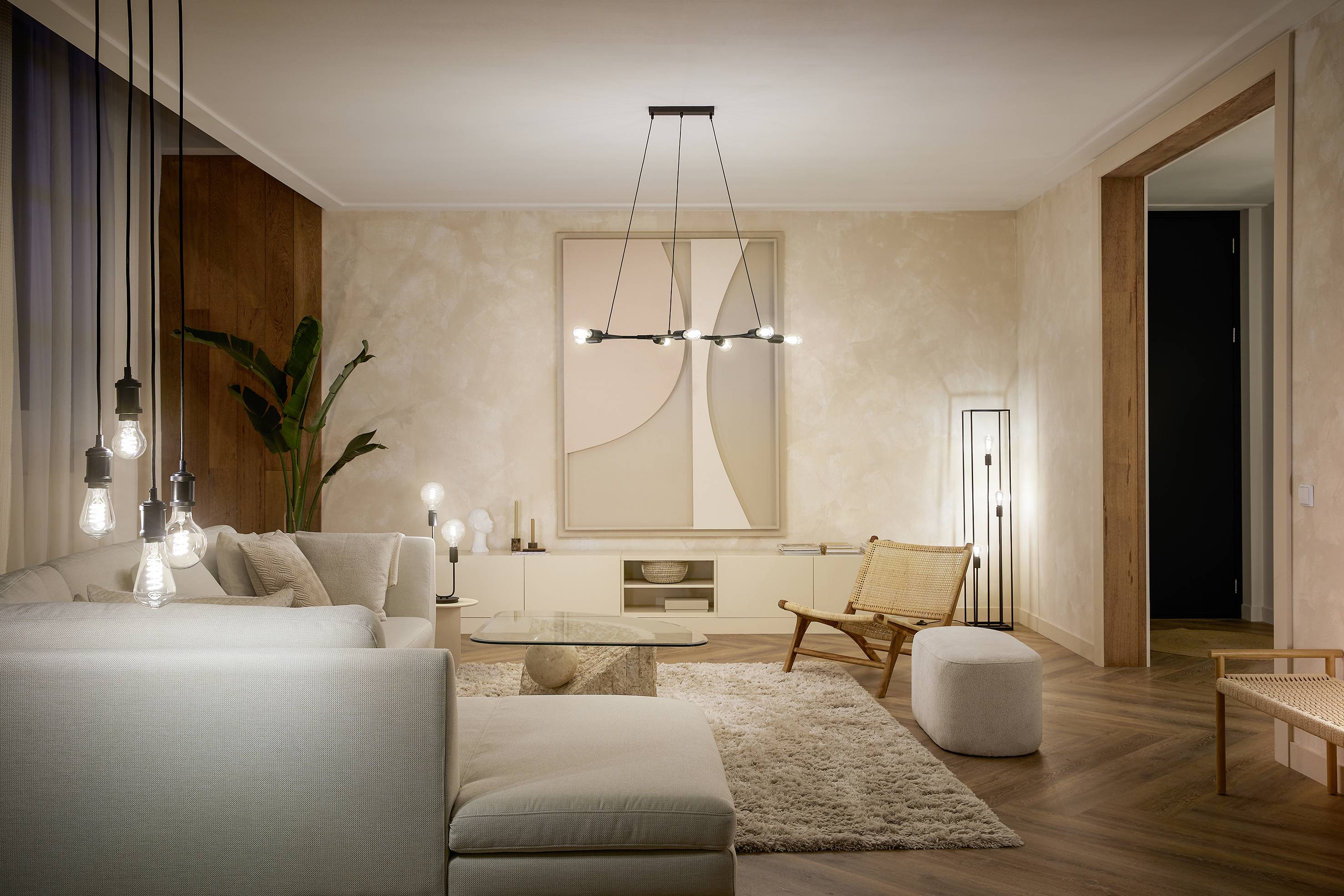 A modern living room with a beige sofa, rug and understated artwork on the wall. Pendant lights and plants create a cosy atmosphere.