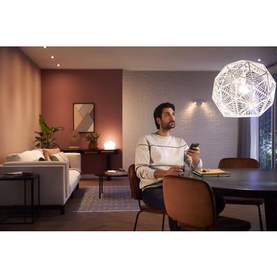 A man sits at a table in a modernly furnished living room, holding a remote control and gazing pensively upwards.